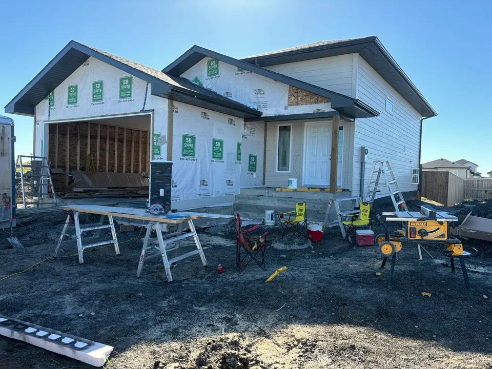 Real job-site photo of a bungalow with an attached garage at the house-wrap stage, with sawhorses, a table saw and folding chairs on the lot