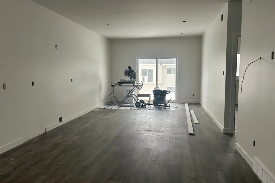 Real job-site photo of an unfinished living room with vinyl plank flooring, bare walls and a mitre saw set up in front of a patio door