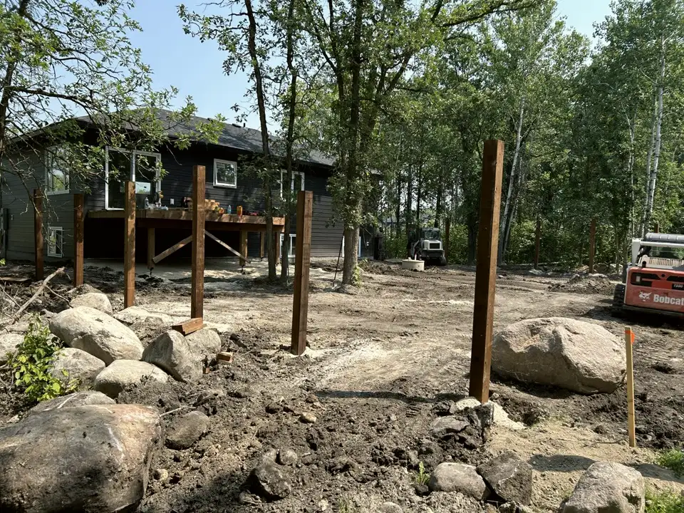 Backyard site work behind a dark-sided home, with wooden posts set in the ground, boulders, a skid steer and bare graded soil among trees
